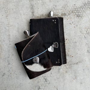 Pode incluir: Dois diários retangulares cobertos com pele de vaca preta e branca. O diário maior apresenta um pingente de coração prateado e um clipe de metal. O diário menor tem um elástico azul claro, um coração prateado e uma aba frontal curva.