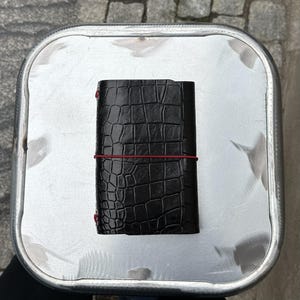 Peut inclure: Un carnet noir avec un motif texturé en peau de crocodile, maintenu par un fin élastique rouge vif qui s'enroule horizontalement autour du centre de la couverture, avec de petits œillets métalliques rouges sur le bord du dos.