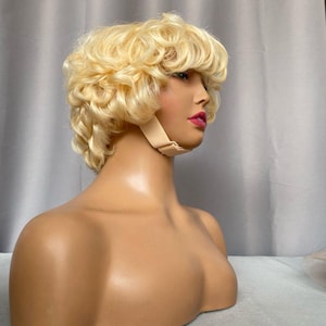 Puede incluir: Peluca rubia con pelo corto y rizado, exhibida en una cabeza de maniquí. La peluca tiene raya a un lado y una línea de cabello de aspecto natural. El maniquí tiene tez bronceada, lápiz labial rosa y una correa beige alrededor de la cabeza.