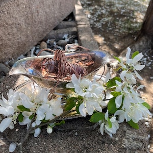 Puede incluir: Una fuente plateada con una escultura de alambre de color cobre en su interior, adornada con flores blancas y hojas verdes. La fuente descansa sobre una superficie de piedra, creando una exhibición natural y elegante.