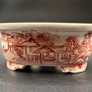 May include: A small, off-white ceramic bonsai pot with a red landscape design. The pot has a rectangular shape with rounded corners and three small feet. The landscape depicts buildings and trees.