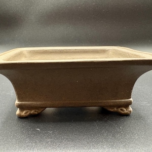 May include: A brown rectangular bonsai pot with a slightly flared shape. The pot has a wide, shallow basin and sits on four small, decorative feet. The overall design is simple and elegant, suitable for showcasing miniature trees.