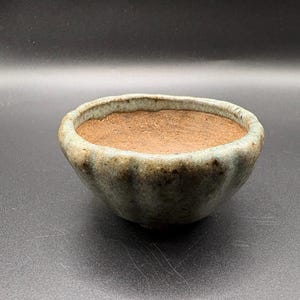 May include: A small, handcrafted ceramic bowl with a textured, light green exterior and a brown interior. The bowl has a rounded shape with a slightly uneven rim. The pottery is likely for decorative or functional use.