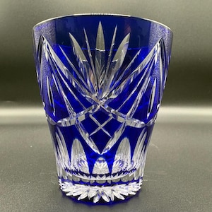 Puede incluir: Un vaso de cristal tallado azul cobalto. El cristal presenta un patrón detallado de cortes verticales y angulados, creando un diseño de estrella. El borde y la base tienen un patrón floral. El vaso mide aproximadamente 10 cm de alto.