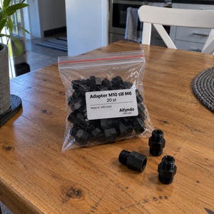 Op de afbeelding: Een doorzichtige, hersluitbare plastic zak met meerdere zwarte schroefdraadadapters, met drie extra adapters buiten de zak. Het etiket op de zak vermeldt Adapter M10 till M6, 20 st, Material: ABS-plast, en Aifyndo Sweden.