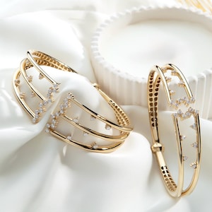 May include: Two gold-toned cuff bracelets featuring a multi-layered design with open gaps. Each bracelet is adorned with small, clear, sparkling stones set along the edges of the metal bands, creating a geometric and layered appearance.