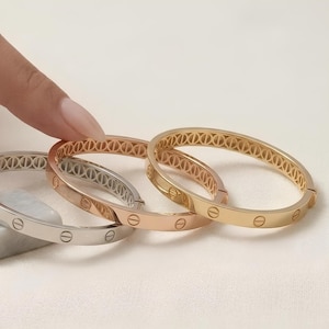 May include: Three metal bangle bracelets in silver, rose gold, and yellow gold tones. Each circular bracelet features a repeating pattern of engraved screw head motifs along the outer surface and a decorative openwork lattice design on the inner band.