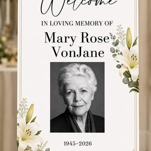 May include: A rectangular memorial sign featuring the text Welcome, In Loving Memory Of, Mary Rose VonJane, and 1945-2026. The design includes a black and white portrait of an older person, framed by white lily flowers and green eucalyptus leaves.
