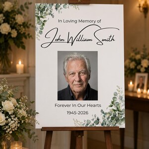 May include: A white memorial sign on a wooden easel featuring the text In Loving Memory of John William Smith, a portrait of an older man, the text Forever In Our Hearts, and the dates 1945-2026, decorated with green eucalyptus leaf illustrations.