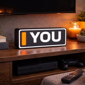 May include: Black rectangular LED sign with the words "I YOU" in white. The "I" is represented by an orange vertical bar. The sign is on a wooden surface with a TV, plants, and other decor in the background.