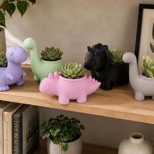 May include: A collection of five dinosaur-shaped planters in purple, light green, pink, black, and grey, each holding a small green succulent plant. The planters feature distinct dinosaur silhouettes with openings on their backs for the plants.