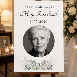 May include: A white memorial sign on a wooden easel featuring the text In Loving Memory Of Mary Rose Smith 1945 2026, a circular black and white portrait of an older person, and a watercolor illustration of green leaves at the bottom.