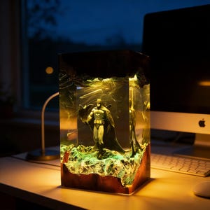 May include: A Batman figurine encased in a rectangular resin lamp. The lamp features a wooden base and a glowing green and yellow interior. The superhero figure is in a classic pose, with a dark cape.
