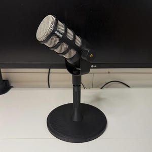 May include: A black RØDE microphone with a silver mesh head is mounted on a black stand. The microphone has a distinctive design with a black body and a silver mesh grille. The stand has a round base and a vertical support.