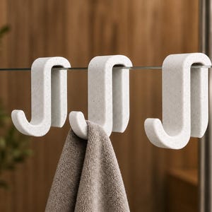 May include: Three white S-shaped hooks with a speckled texture hanging over a thin horizontal glass edge. The hooks are made of a solid material and feature a curved design for hanging items, with a grey towel draped over the center hook.