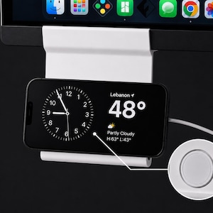 May include: A white metal mount attached to a computer monitor, holding a smartphone in landscape orientation. The phone screen displays an analog clock, the location Lebanon, and a temperature of 48 degrees with partly cloudy weather.