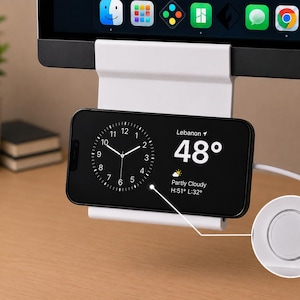 May include: A white monitor-mounted smartphone holder supporting a black smartphone in landscape orientation. The phone screen displays an analog clock, the location Lebanon, 48 degrees, and weather conditions. A circular magnetic charger is visible.