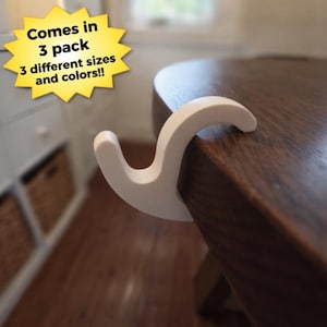 May include: A white, curved table hook is attached to the edge of a wooden table. The hook is designed to hang items. Text on a yellow starburst reads "Comes in 3 pack 3 different sizes and colors!!"