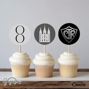 May include: Three white cupcakes with black and silver themed cupcake toppers. The toppers feature a number 8, a temple, and the letters CTR.