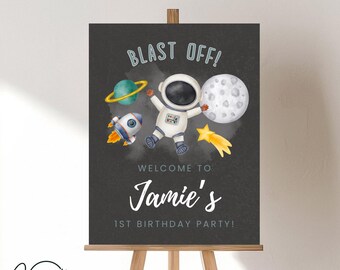 May include: A black chalkboard sign with a white astronaut, rocket, planets, and stars. The sign says "Blast Off! Welcome to Jamie's 1st Birthday Party!"