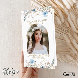 Puede incluir: Una tarjeta blanca con diseños florales azules y dorados. La tarjeta dice "Bienvenido al Bautismo de Isabella Williams 23 de abril de 20xx". Una foto de una niña con un vestido blanco está en el centro de la tarjeta.