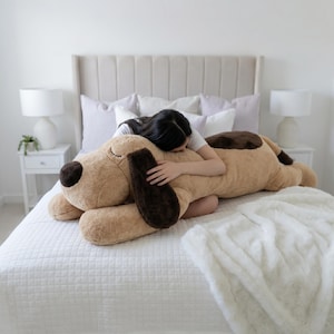 May include: A large, tan plush dog toy with long, dark brown ears, a dark brown nose, and a dark brown spot on its back. The stuffed animal features embroidered closed eyes and a soft, fuzzy texture, designed in a long, reclining position.