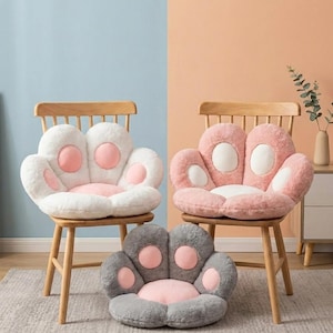 May include: Three plush paw-shaped seat cushions in white, pink, and gray, each with pink accents. The cushions are on wooden chairs, with one cushion on the floor. The background is a mix of blue and peach.