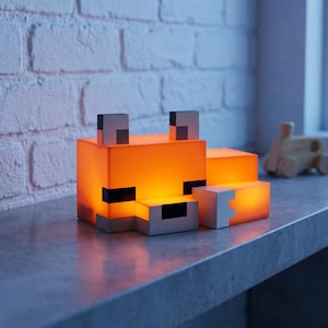 May include: An orange and white fox-shaped lamp with black accents. The lamp is illuminated from within, creating a warm glow. The design is reminiscent of a pixelated video game character. The lamp is sitting on a gray surface.