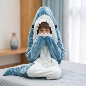 May include: A blue shark onesie with a white belly and a hood featuring a shark's face. The onesie has a zipper closure and is made of soft, plush material. The shark's face includes white teeth and black eyes.
