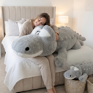 May include: A large, grey alligator soft toy with white details and black eyes. The alligator is on a bed. A smaller alligator plush is in a wicker basket. The toy is soft and cuddly, perfect for children.