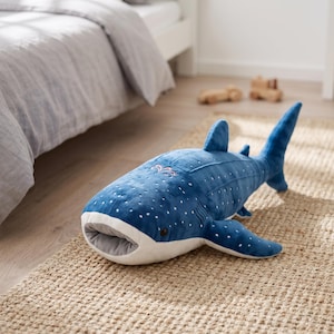 May include: A plush whale shark toy featuring a deep blue body with white polka dot patterns, a wide light grey mouth, dark eyes, and prominent fins. The soft fabric toy has a rounded head and a long, tapering tail section with a large fin.