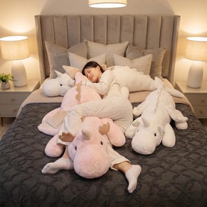 May include: A person is asleep on a bed with large plush unicorn pillows in white and pink. The bed has a grey comforter and several decorative pillows. Two lamps with beige shades are on either side of the bed.