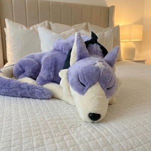 May include: A lavender and white plush wolf toy with closed eyes, lying on a white bed. The toy has a black bow and is designed to resemble a sleeping wolf. The soft toy is a decorative item.