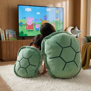 May include: Two plush turtle shells in a soft sage green color, featuring dark green embroidered hexagonal patterns on the back. These wearable, rounded cushions are designed to be worn on the back, mimicking the appearance of a turtle shell.