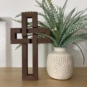 May include: A dark brown wooden cross with a unique interlocking design stands on a light brown shelf. Beside it is a cream-colored vase with a textured surface, filled with green and gray faux greenery. The cross is a decorative religious item.