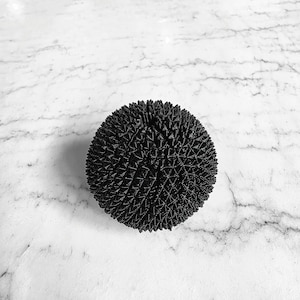 May include: A black, spherical object with a textured, spiky surface. The object is centered in the frame against a white marble background. The spiky texture creates a geometric pattern across the sphere's surface.