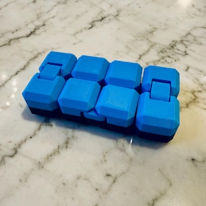 Può includere: Un giocattolo fidget blu e nero con otto blocchi quadrati collegati. I blocchi sono di un vibrante colore blu e sono collegati da cerniere, che consentono loro di muoversi e piegarsi. La base del giocattolo è nera.