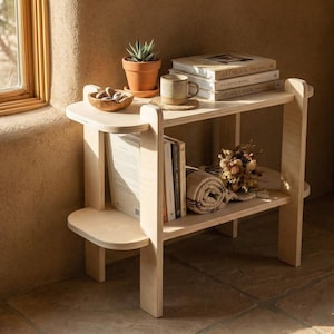 Può includere: Uno scaffale in legno chiaro a due livelli con bordi arrotondati. Il ripiano superiore ospita un vaso in terracotta con una pianta grassa, una piccola ciotola in legno con pietre e una tazza su un sottobicchiere. Il ripiano inferiore contiene tre libri e un tessuto arrotolato.