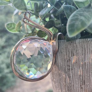May include: A clear, multi-faceted spherical crystal ornament encased in a curved, metallic silver-toned wire frame. The circular crystal features numerous triangular cuts that refract light, creating subtle rainbow reflections across its surface.