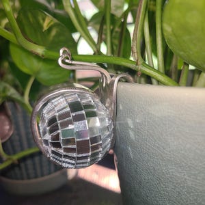 May include: A decorative snail-shaped ornament crafted from a silver-toned metal fork, featuring a spherical disco ball made of small, reflective square mirror tiles as its shell, designed to hook onto the edge of a plant pot or container.