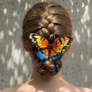 Puede incluir: Un accesorio para el cabello con forma de mariposa con alas de colores naranja, amarillo y azul vibrantes, sujeto a una pinza para el cabello marr&oacute;n. La mariposa se coloca sobre un peinado de mo&ntilde;o trenzado, mostrando patrones de alas detallados.