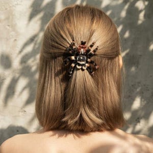 May include: A hair clip shaped like a tarantula spider with black, white, and reddish-brown fuzzy textures. The spider is positioned at the centre of the back of the head, securing a section of light brown hair in a half-up hairstyle.