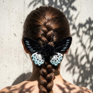 May include: A hair accessory shaped like a butterfly with a black velvet body and wings. The upper wings are solid black, while the lower wings feature a light blue and white gradient with black spots. The butterfly is attached to brown braided hair.