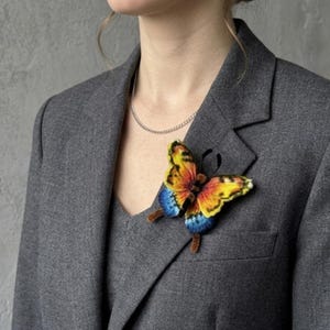 Puede incluir: Un broche de mariposa con alas vibrantes en amarillo, naranja y azul con marcas negras, prendido en un blazer gris. La mariposa tiene un cuerpo marr&oacute;n oscuro y difuso y antenas negras delgadas, creando una pieza de accesorio detallada.