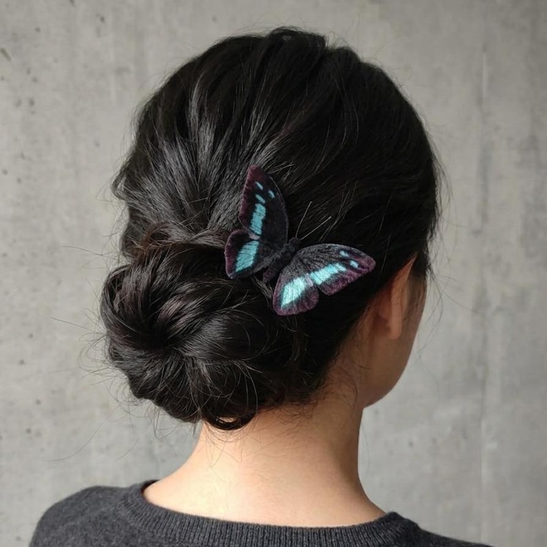 May include: A black butterfly hair clip with teal and purple accents is pinned into dark hair styled in a low bun. The butterfly's wings are detailed with intricate patterns, adding a touch of elegance to the hairstyle.