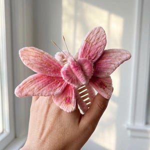 May include: A pink butterfly hair claw clip featuring a soft, fuzzy velvet-like texture on the wings. The wings are detailed with delicate gold embroidered veins and thin, dark antennae. The base of the clip is a standard plastic hair claw.