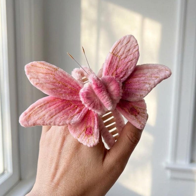 May include: A pink butterfly hair claw clip featuring a soft, fuzzy velvet-like texture on the wings. The wings are detailed with delicate gold embroidered veins and thin, dark antennae. The base of the clip is a standard plastic hair claw.