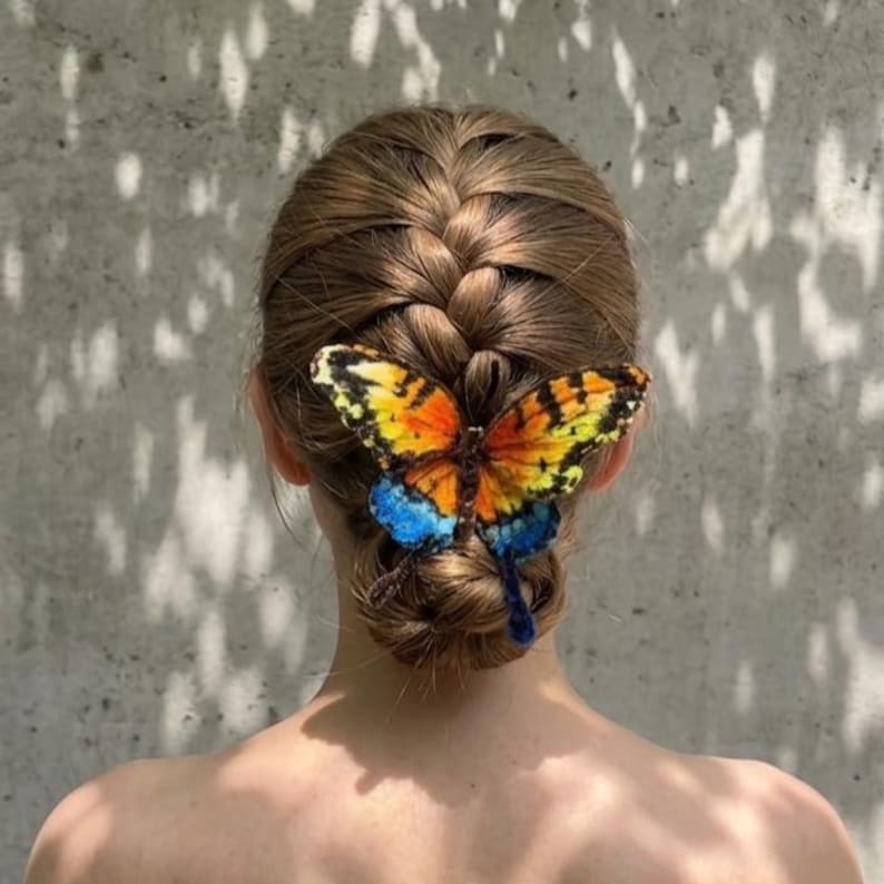 Puede incluir: Un accesorio para el cabello con forma de mariposa con alas de colores naranja, amarillo y azul vibrantes, sujeto a una pinza para el cabello marr&oacute;n. La mariposa se coloca sobre un peinado de mo&ntilde;o trenzado, mostrando patrones de alas detallados.