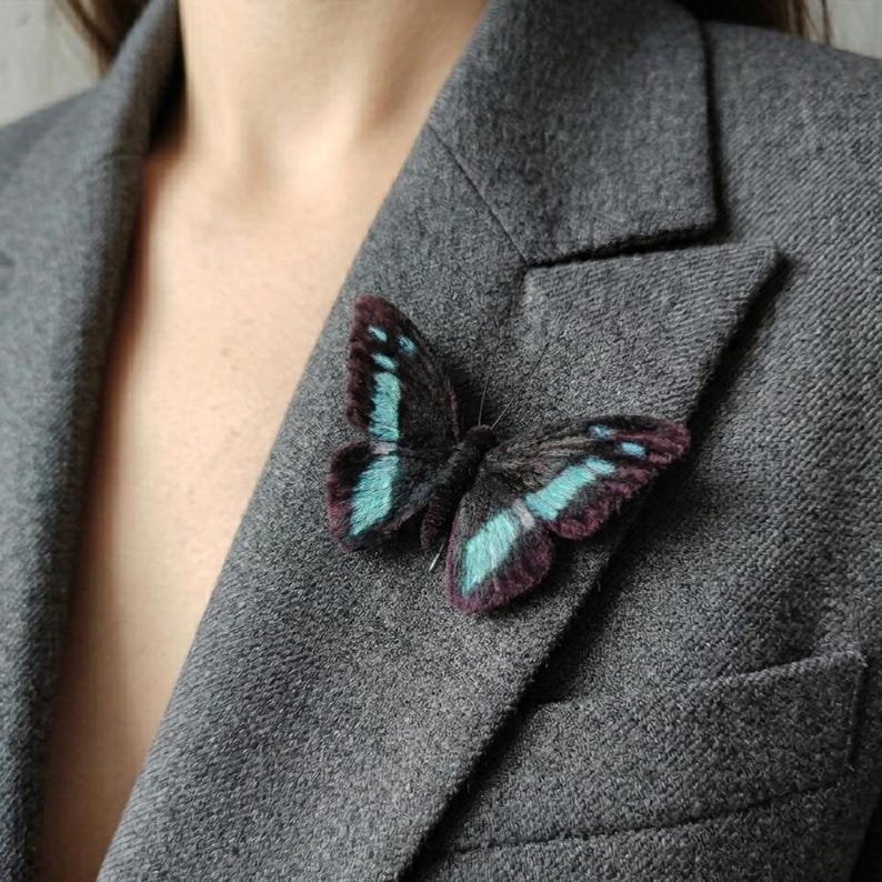 May include: A butterfly brooch with teal and purple accents is pinned to a gray blazer. The butterfly's wings are detailed with teal stripes and purple edges, creating a striking contrast against the dark body.