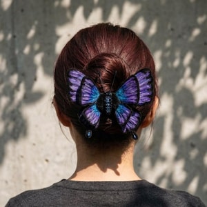 May include: A purple and blue butterfly hair clip is attached to a person's dark brown hair, styled in a bun. The butterfly has intricate details and a beaded body. Two blue teardrop-shaped beads dangle below the butterfly.
